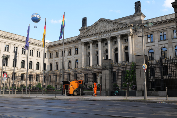 Large modern building with many windows, identified as the Bundestag in Berlin, Germany, surrounded by street infrastructure, pedestrians, trees, and a cloudy sky.