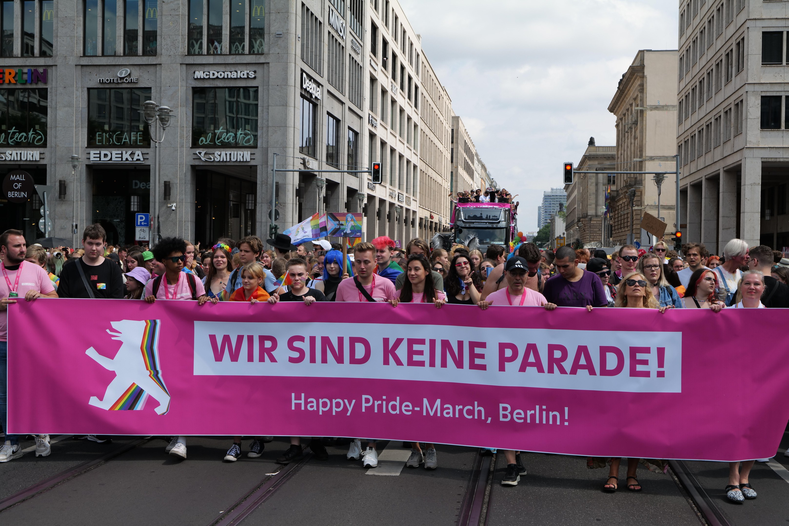 Eine Gruppe von Menschen marschiert auf einer Straße in Berlin, Deutschland, mit einer pinken Fahne mit der Aufschrift "Happy Pride March", umgeben von Gebäuden, Laternenmasten und Verkehrszeichen unter einem bewölkten Himmel.