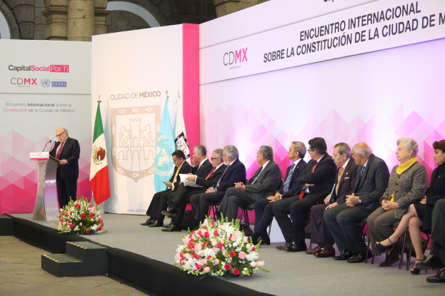 Gruppe von Menschen, die auf Stühlen sitzen und einem Podium mit Mikrofon gegenüberstehen, umgeben von Blumenarrangements, Flaggen und Texttafeln in einem formellen Saal mit Säulen und einer Wand.