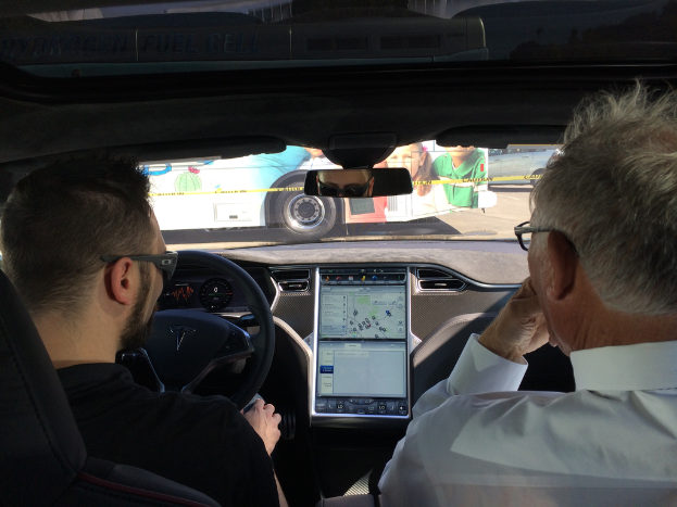 Eine Person fährt ein Auto mit einer Routenkarte neben dem Lenkrad, während eine andere Person auf dem Vordersitz sitzt; ein Bus fährt vor dem Auto auf der Straße vorbei.