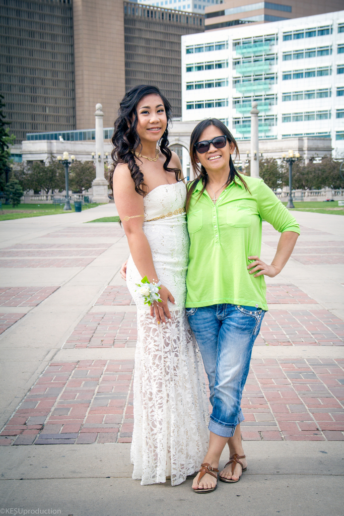 Zwei Frauen auf einem Weg, die lächeln, wobei eine ein Kleid trägt und die andere ein Hemd und Brille, mit Gebäuden, Laternen, einem Zaun, Denkmälern und Text in der linken unteren Ecke und Gras im Hintergrund.