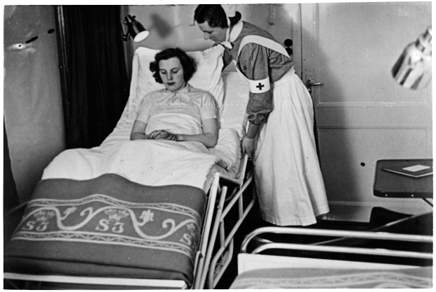 Ein altes Schwarz-Weiß-Foto einer Frau in einem Krankenhausbett mit einer Krankenschwester daneben, einem Tisch mit verschiedenen Gegenständen rechts daneben und einem Vorhang und einer Wand im Hintergrund.