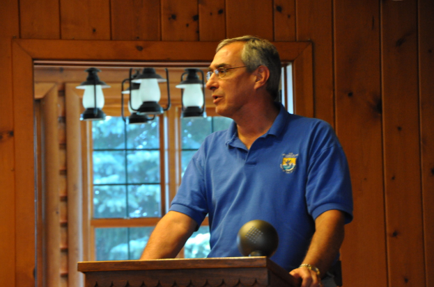 Eine Person in einem blauen T-Shirt steht hinter einem hölzernen Podium im Innern, mit einer Wand, einem Fenster und hängenden Lampen im Hintergrund.