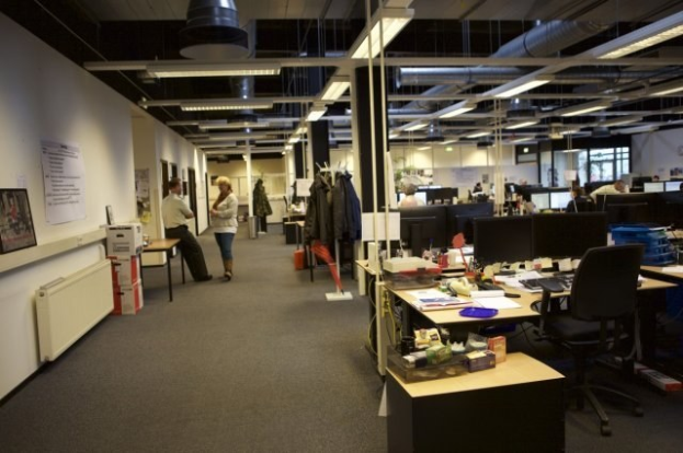 Großes Großraumbüro mit Menschen an Schreibtischen, Computern und Monitoren; Papiere, Bücher und Kartons auf Tischen; Kleiderbügel, Tafeln mit Text und Bildern an den Wänden und Deckenlampen.