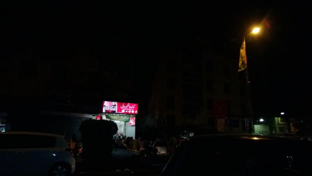 Eine nächtliche Szenerie einer Straße mit Autos, einem kleinen Geschäft im Hintergrund, einem Laternenmast auf der rechten Seite, einer Reklamewand über dem Geschäft und beleuchteten Gebäuden auf der rechten Seite.