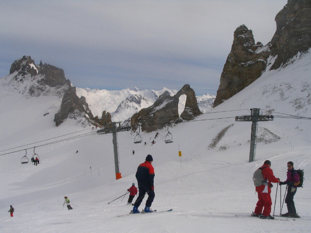Menschen in Pullovern fahren Ski auf Eis, mit Seilbahn, Bergen und bewölktem Himmel im Hintergrund.