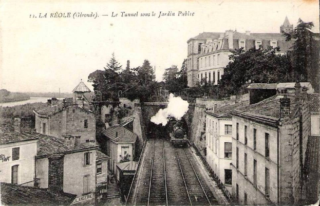 Black and white photograph of a train on tracks passing through a tunnel labeled "la réole gironde - le tunnel sous le jardin public," with buildings, trees, and water visible around it.