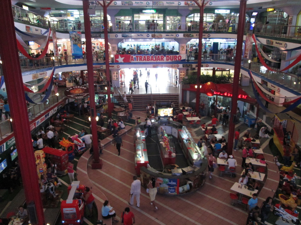 Eine belebte Einkaufsmeile mit Säulen, Menschen, Tischen, Fliesenboden, Flaggen, Geschäften, einem Banner, Treppen und einem Namensschild.