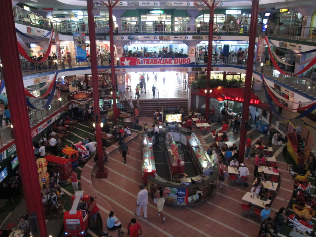 Eine belebte Einkaufsmeile mit Säulen, Menschen, Tischen, Fliesenboden, Flaggen, Geschäften, einem Banner, Treppen und einem Namensschild.