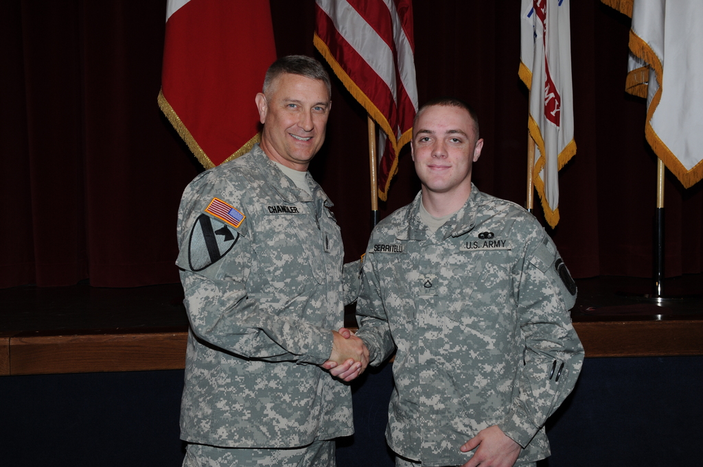 Zwei Personen in Uniformen geben sich die Hand und lächeln, vor vier Fahnen an Stangen gegen einen dunklen Hintergrund.