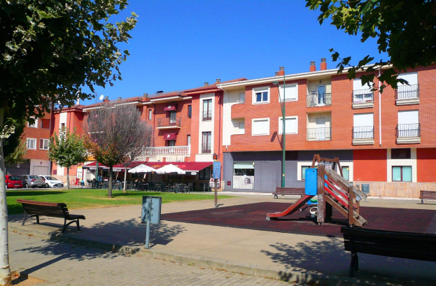 Ein Park mit einem Spielplatz, umgeben von Gebäuden mit Bänken, Bäumen, Gras, einem Schild, Fahrzeugen, Zelten, Menschen unter einem Dach und einer bewölkten Himmel.