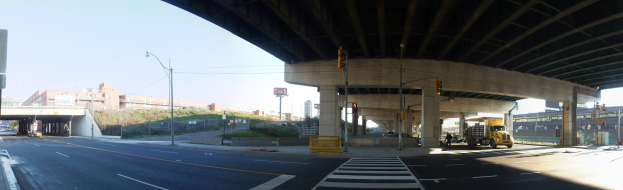 A truck driving under a bridge on a city street with buildings, street infrastructure, and vehicles under a clear blue sky.