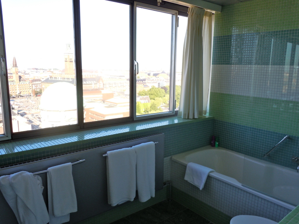 Ein Badezimmer mit einer Badewanne, Handtüchern, Fenstern mit Vorhängen und einem Blick auf Gebäude, Bäume und den Himmel.