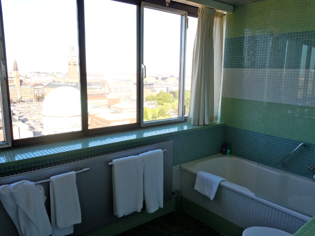 Ein Badezimmer mit einer Badewanne, Handtüchern, Fenstern mit Vorhängen und einem Blick auf Gebäude, Bäume und den Himmel.