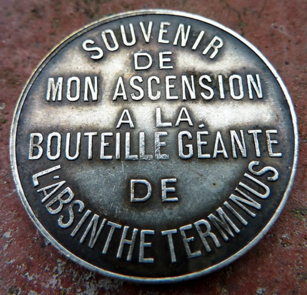 Round metal plaque on a building with the inscription "Souvenir de Mon Ascension à la Bouteille Gente de L'Absinthe Terminus".