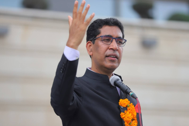 A man in a black suit and tie, wearing spectacles and a garland, speaks into a microphone during a speech with a blurred background.