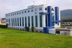 Großes weißes und blaues Gebäude mit Fahrzeugen, Straßeninfrastruktur und Bergen im Hintergrund, auf einer grünen Wiese mit Bäumen und Gras gelegen.