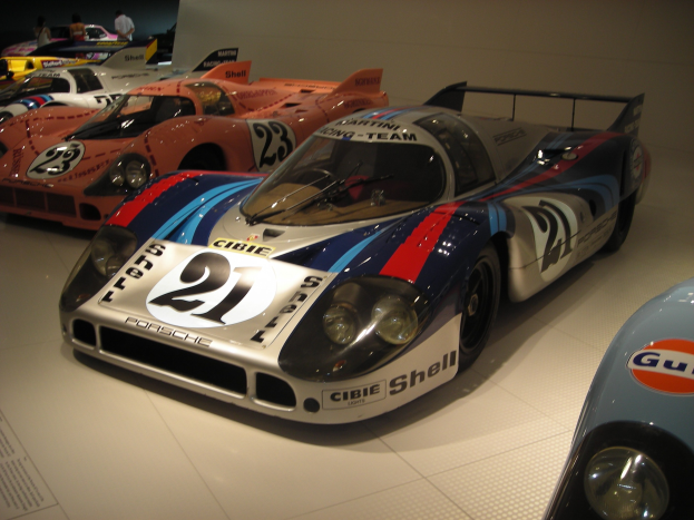 Ein Porsche 917K Le Mans Rennauto in einem Museum neben anderen Sportwagen ausgestellt, mit Menschen, die es beobachten, und einem Textschild auf der linken Seite.