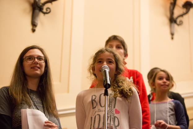 Eine Gruppe junger Mädchen steht Seite an Seite vor einem Mikrofon, eine hält ein Blatt Papier in der Hand, mit an der Wand angebrachten Lampen im Hintergrund, die eine warme Atmosphäre schaffen.