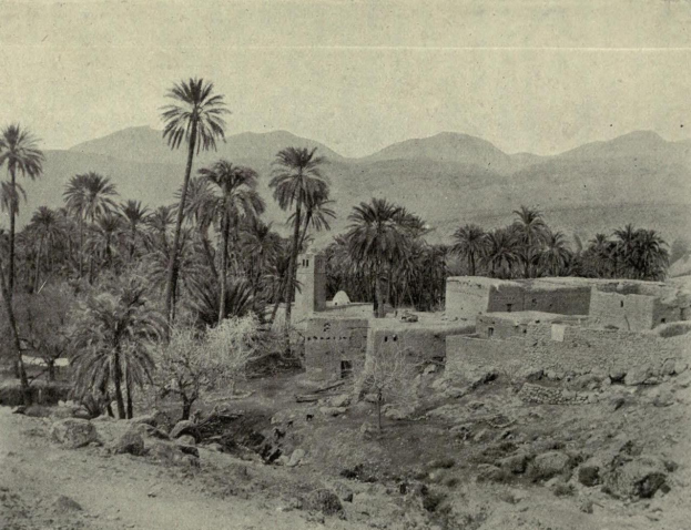 Old black and white photo of a desert village with houses, palm trees, vegetation, rocks, and mountains in the background, featuring text at the bottom.