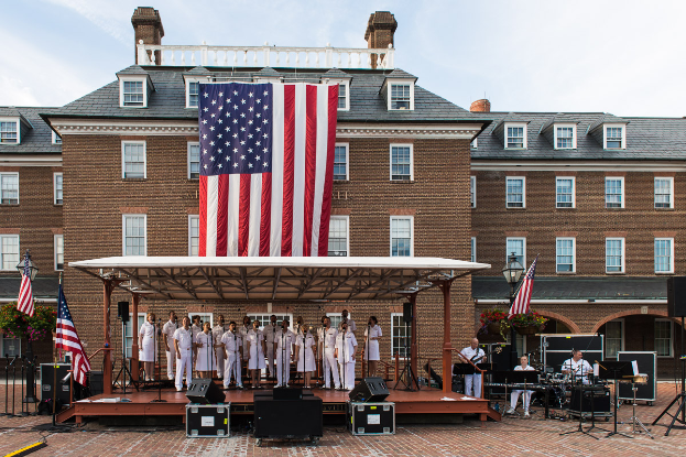 Eine Gruppe von Menschen auf einer Bühne vor einem Gebäude mit Fenstern, umgeben von Fahnen, Lautsprechern und Blumenpflanzen, die Musikinstrumente spielen, mit Laternen und einem bewölkten Himmel im Hintergrund, was auf ein Gedenkkonzert auf dem Mount Vernon hindeutet.