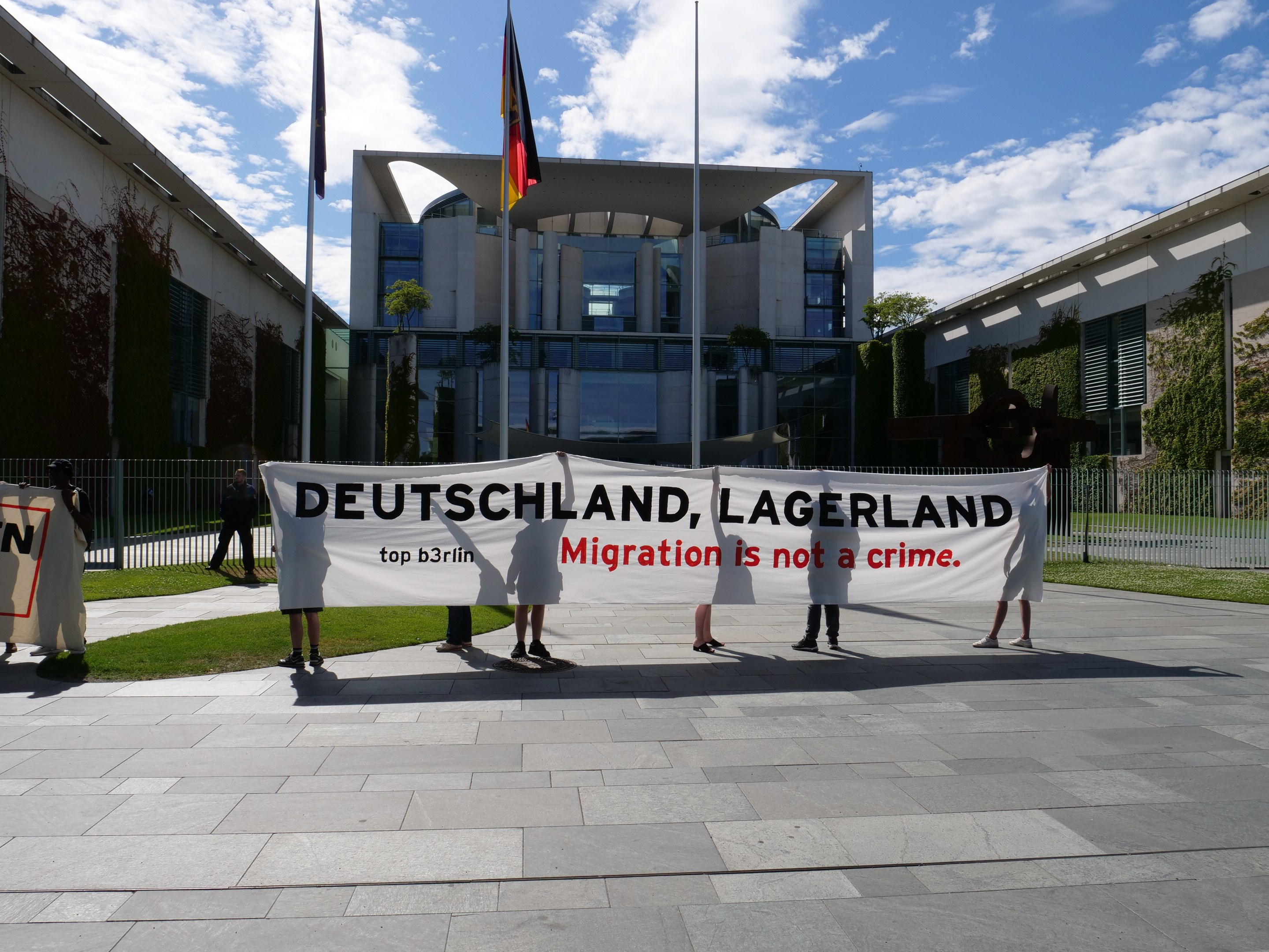 Group of people holding a banner reading "Deutschland, Lagerland Migration is Not a Crime" in front of a building with windows, surrounded by grass, a metal fence, plants, trees, and flags with poles under a cloudy sky.