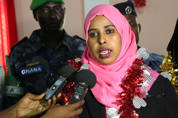 Frau mit pinkem Kopftuch spricht mit Reportern während eines Interviews, wobei ein Mikrofon links und eine Vorhangwand im Hintergrund zu sehen ist.