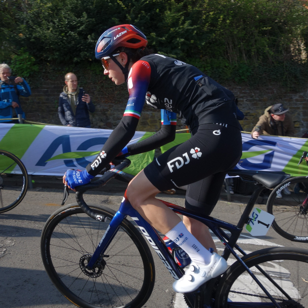 Die professionelle Radrennfahrerin Katie Archibald fährt ein Fahrrad die Straße entlang, trägt einen Helm, eine Brille, Handschuhe und Schuhe und hat eine Menschenmenge neben sich.