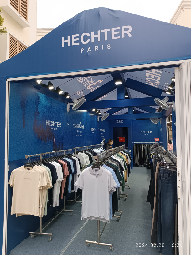 Ein Hechter Paris Stand auf der Paris Fashion Week, mit Kleidung an Bügeln, einer Person im Stand, Lichtern, einem Gebäude im Hintergrund und einem Baum auf der linken Seite, mit einem sichtbaren Himmel oben und einem Wasserzeichen in der rechten unteren Ecke.