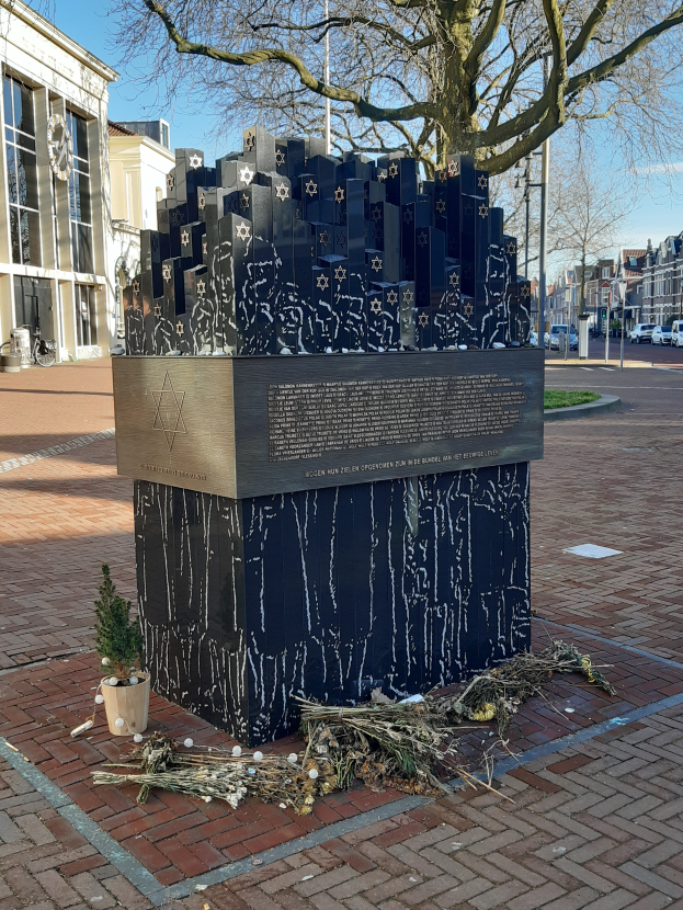 Ein Denkmal für Holocaust-Opfer steht in einem Stadtplatz mit einem Baum davor, einem Topf mit einer Pflanze und verstreuten trockenen Blättern, Gebäuden, Fahrzeugen, Pfosten und Bäumen im Hintergrund.