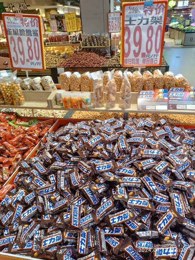 Vitrine voller verschiedener Süßigkeiten, darunter Snickers, in einem Geschäft mit Lebensmittelartikeln auf Regalen, Texttafeln, Deckenleuchten und Hintergrundobjekten.
