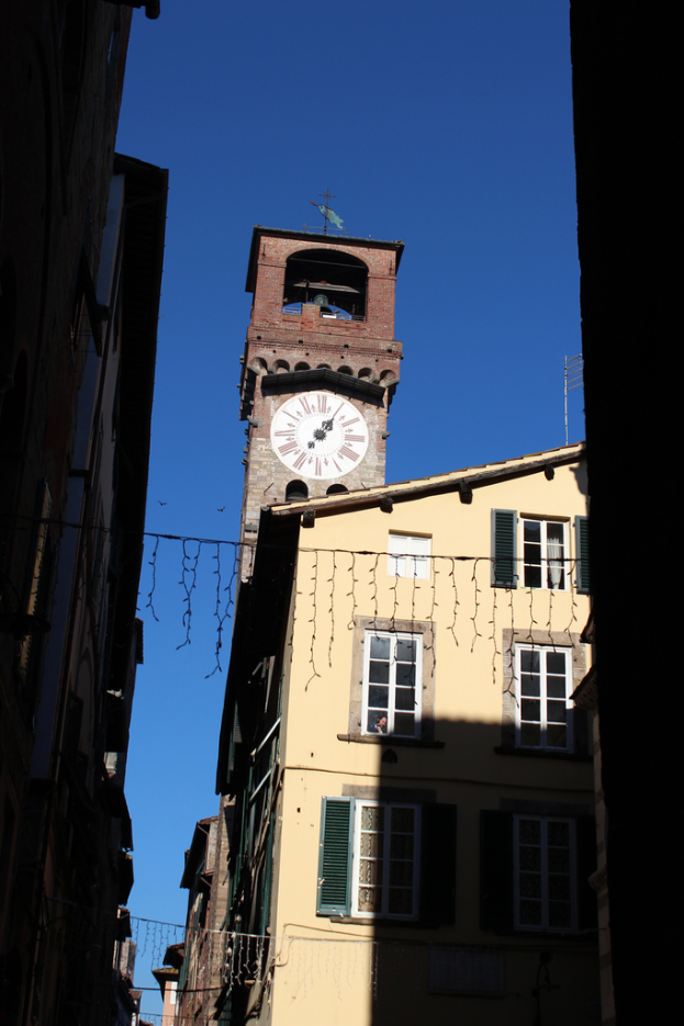 Ein Uhrenturm mit einer Antenne an seiner Spitze, umgeben von Gebäuden mit Glasfenstern auf der rechten Seite, unter einem blauen Himmel.