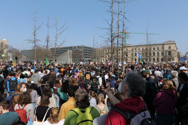 Eine große Menge von Menschen, die vor einem Gebäude mit Fenstern, Bäumen und einem klaren Himmel im Hintergrund stehen, viele tragen Taschen und halten Schilder.