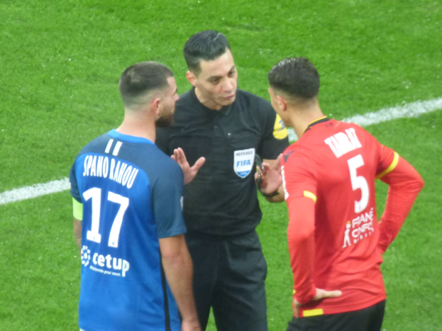 Schiedsrichter in schwarzem Hemd und Hose, der mit zwei Männern auf einem Fußballfeld spricht, einer in blauen und roten T-Shirts mit Text und Brille, der andere mit Brille.