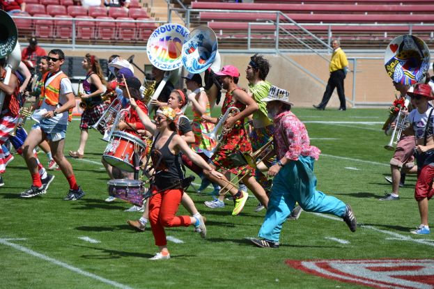 Eine Gruppe von Menschen versammelt sich im Freien mit musikalischen Trommeln, verschiedenen Instrumenten, Stühlen, Treppen und einem Zaun im Hintergrund.