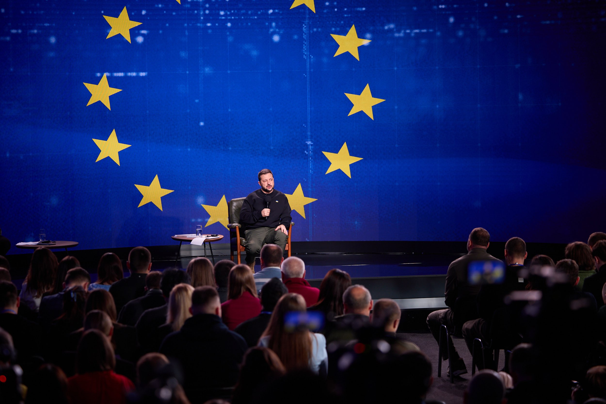 A man in a black dress sits on a stage holding a microphone, speaking to an audience seated in chairs, with two tables and a blue screen displaying yellow stars in the background.