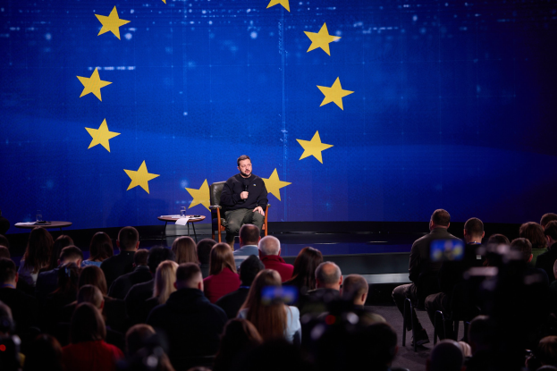 A man in a black dress sits on a stage holding a microphone, speaking to an audience seated in chairs, with two tables and a blue screen displaying yellow stars in the background.