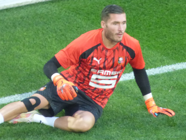 Ein Fussballspieler in rotem T-Shirt, schwarzen Shorts, weissen Socken und orangen Baseballhandschuhen sitzt auf dem Boden mit den Händen auf den Knien.
