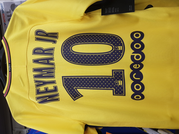 Ein gelbes Fußballtrikot mit der Nummer 10, das Neymar Jr. von der Heimkit des PSG 2018-19 gehört, wird mit einem schwarzen Etikett und Papieren und anderen Gegenständen im Hintergrund gezeigt.