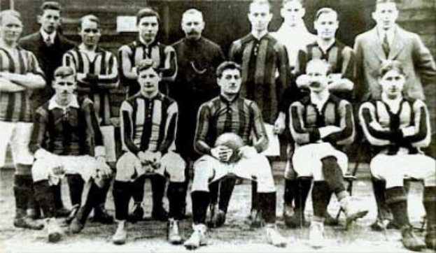 Black and white photo of a soccer team, some standing and some sitting, in front of a wall with "Ballymena Football Club" text at the bottom.