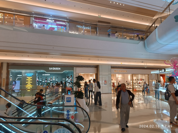 Interior of a shopping mall with people walking, escalators, illuminated ceiling lights, storefronts with name boards, text displays, potted plants, and a visible watermark noting the mall's recent reopening post-coronavirus pandemic.