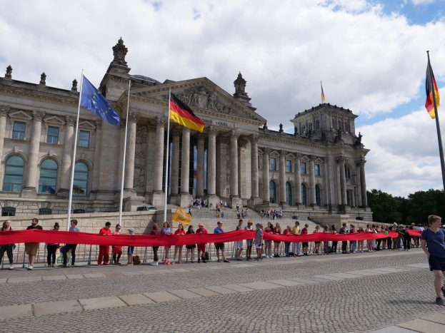 Eine Gruppe von Menschen steht vor dem Reichstagsgebäude in Berlin, Deutschland, das mit Fenstern, Säulen, Bögen und Statuen verziert ist und von Fahnenmasten umgeben ist, mit einem roten Band im Vordergrund, einer Person, die rechts geht, und Bäumen und einem bewölkten Himmel im Hintergrund.