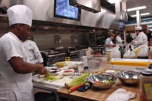 Eine Gruppe von Köchen in einer Küche, die Essen auf einem Schneidbrett zubereiten, mit einem Koch im Vordergrund in einem weißen Kleid und einer Mütze, der ein Messer hält und von verschiedenen Utensilien, Schüsseln, Gemüse, einem Mobiltelefon, Flaschen und anderen Gegenständen umgeben ist, mit einem Fernseher und Deckenleuchten im Hintergrund.