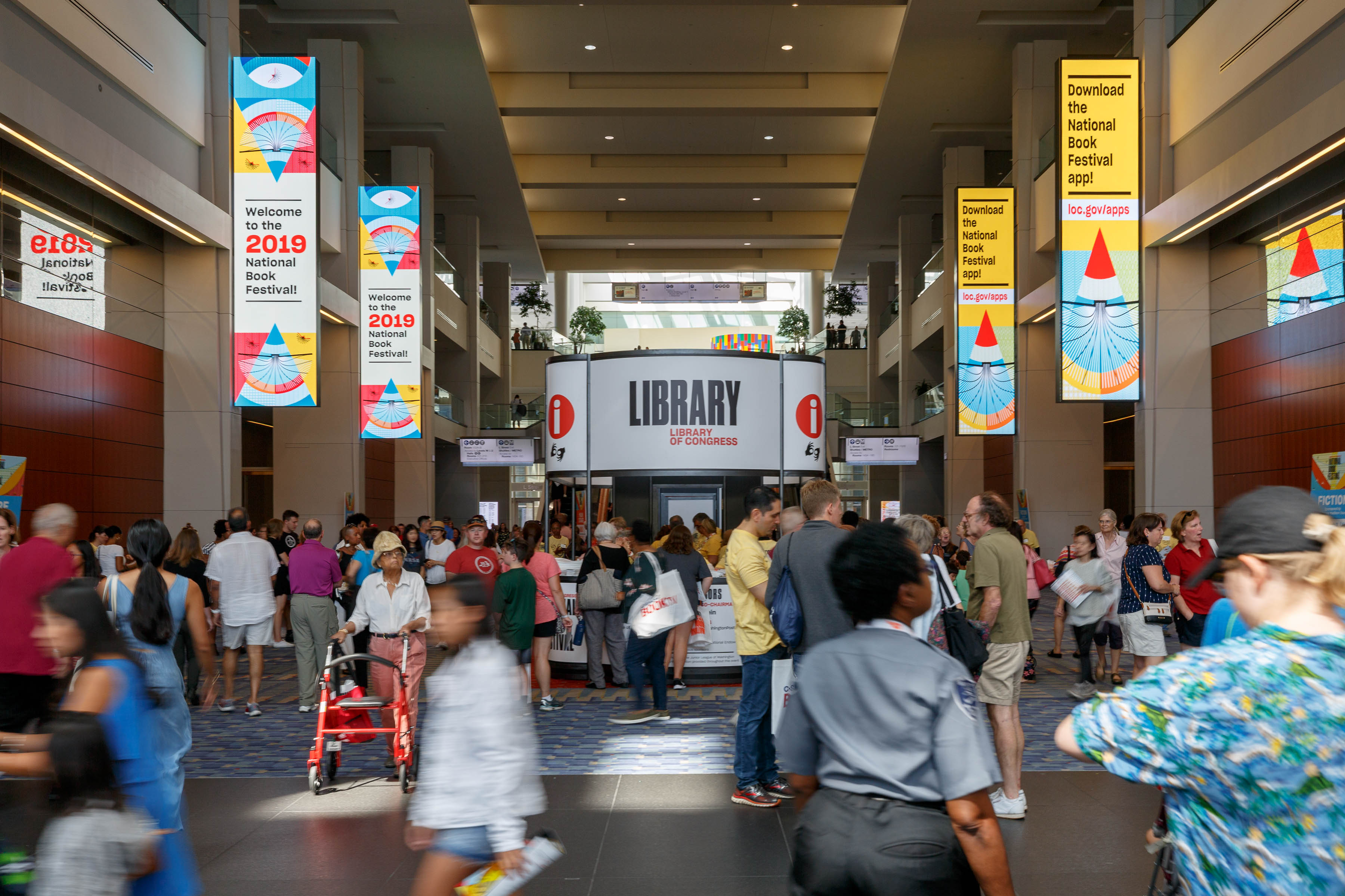 Eine große Gruppe von Menschen, die durch ein Gebäude gehen, einige tragen Taschen und einer schiebt einen Kinderwagen, mit Schildern an Wänden, Säulen und Deckenleuchten und einer Tafel mit Text im Hintergrund.