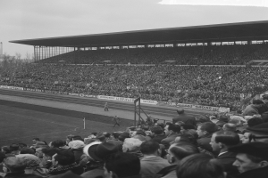 Schwarzes und weißes Foto eines vollen Fußballstadions mit Zuschauern, die stehen und sitzen, Bannern, Pfählen, einem Schuppen, Bäumen, einem Turm und einem bewölkten Himmel.