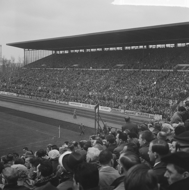 Schwarzes und weißes Foto eines vollen Fußballstadions mit Zuschauern, die stehen und sitzen, Bannern, Pfählen, einem Schuppen, Bäumen, einem Turm und einem bewölkten Himmel.