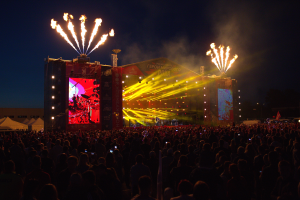 Eine große Menschenmenge versammelte sich auf einem Musikfestival, mit einer Bühne in der Mitte, auf der Musiker Instrumente spielen, beleuchtet von hellen Lichtern und umgeben von Bannern, Zelten links und einem dunklen Himmel mit Feuerwerk im Hintergrund.