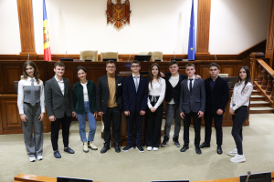 Gruppe von strahlenden Menschen, die vor einem Gerichtssaal mit Monitoren und Mikrofonen auf Tischen, einer hölzernen Plattform mit Stühlen, Flaggen und einem an der Wand montierten Objekt im Hintergrund und einer Treppe mit Geländern auf der rechten Seite posieren.