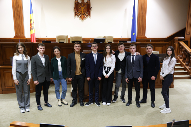 Gruppe von strahlenden Menschen, die vor einem Gerichtssaal mit Monitoren und Mikrofonen auf Tischen, einer hölzernen Plattform mit Stühlen, Flaggen und einem an der Wand montierten Objekt im Hintergrund und einer Treppe mit Geländern auf der rechten Seite posieren.