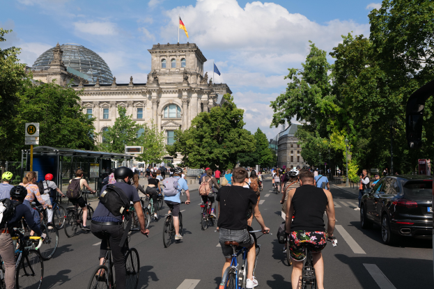 Eine Gruppe von Menschen, die Fahrräder auf einer von Bäumen gesäumten Straße in Berlin, Deutschland, fahren, mit Gebäuden auf beiden Seiten, einem Bushaltestellen auf der rechten Seite und einer Flagge, die auf einem der Gebäude unter einem bewölkten Himmel weht.